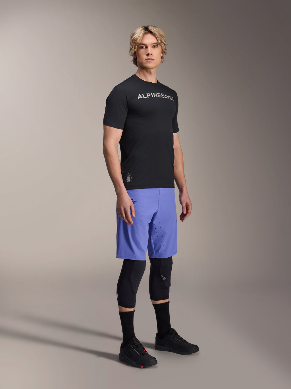 Männliches Model in Alpinestars Mountainbike-Bekleidung, bestehend aus dem schwarzen A-Aria Switch SS Jersey und den A-Aria Lite Shorts in Baja Blue, kombiniert mit schwarzen Knieschützern, schwarzen Socken und schwarzen Mountainbike-Schuhen in einer Ganzkörper-Studioaufnahme.