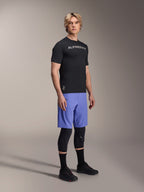 Männliches Model in Alpinestars Mountainbike-Bekleidung, bestehend aus dem schwarzen A-Aria Switch SS Jersey und den A-Aria Lite Shorts in Baja Blue, kombiniert mit schwarzen Knieschützern, schwarzen Socken und schwarzen Mountainbike-Schuhen in einer Ganzkörper-Studioaufnahme.