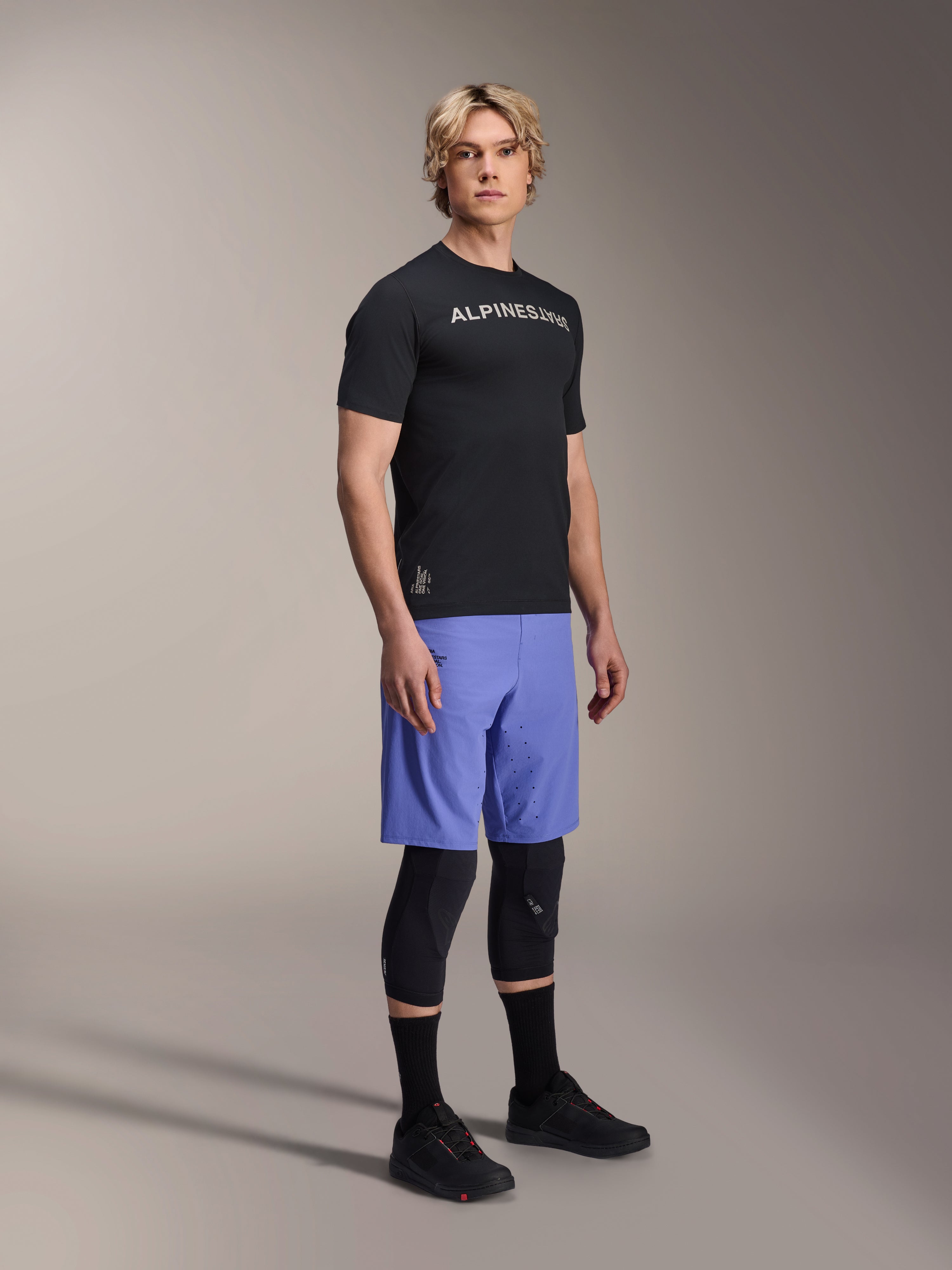 Männliches Model trägt das Alpinestars A-Aria Switch Kurzarmtrikot in Schwarz mit weißem Branding auf der Brust, kombiniert mit hellblauen Mountainbike-Shorts, schwarzen Knieschonern und schwarzen Radschuhen, Ganzkörper-Studioaufnahme für Performance-MTB-Ausrüstung