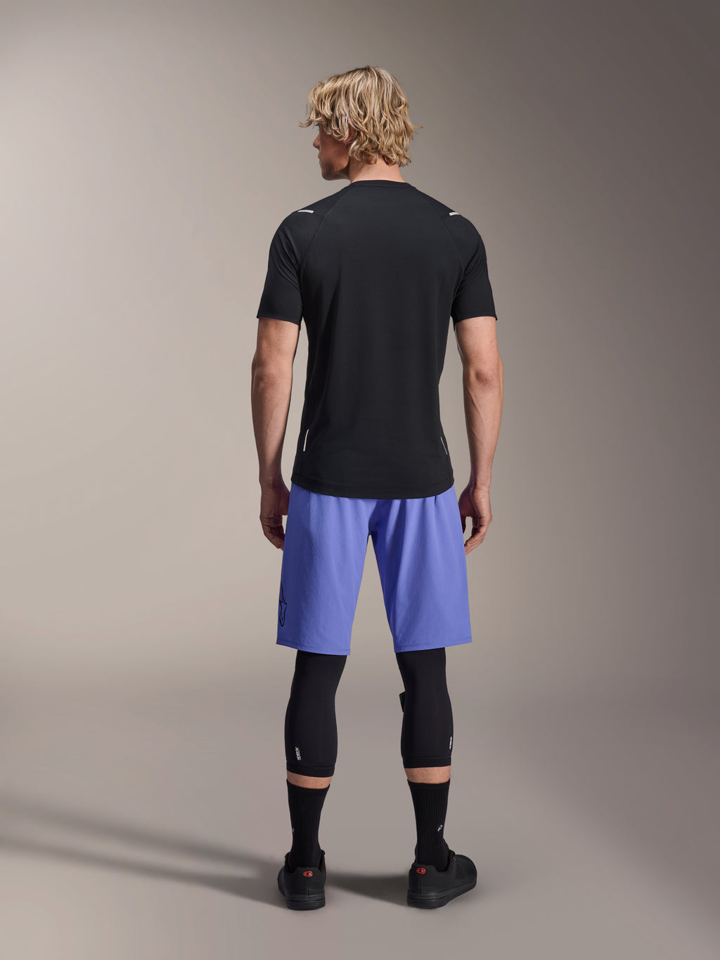 Alpinestars A-Aria Switch Kurzarmtrikot, schwarzes MTB-Trikot, Rückansicht, kombiniert mit blauen Mountainbike-Shorts und schwarzem Beinschutz, Performance-Fahrradausrüstung für Trailfahrten