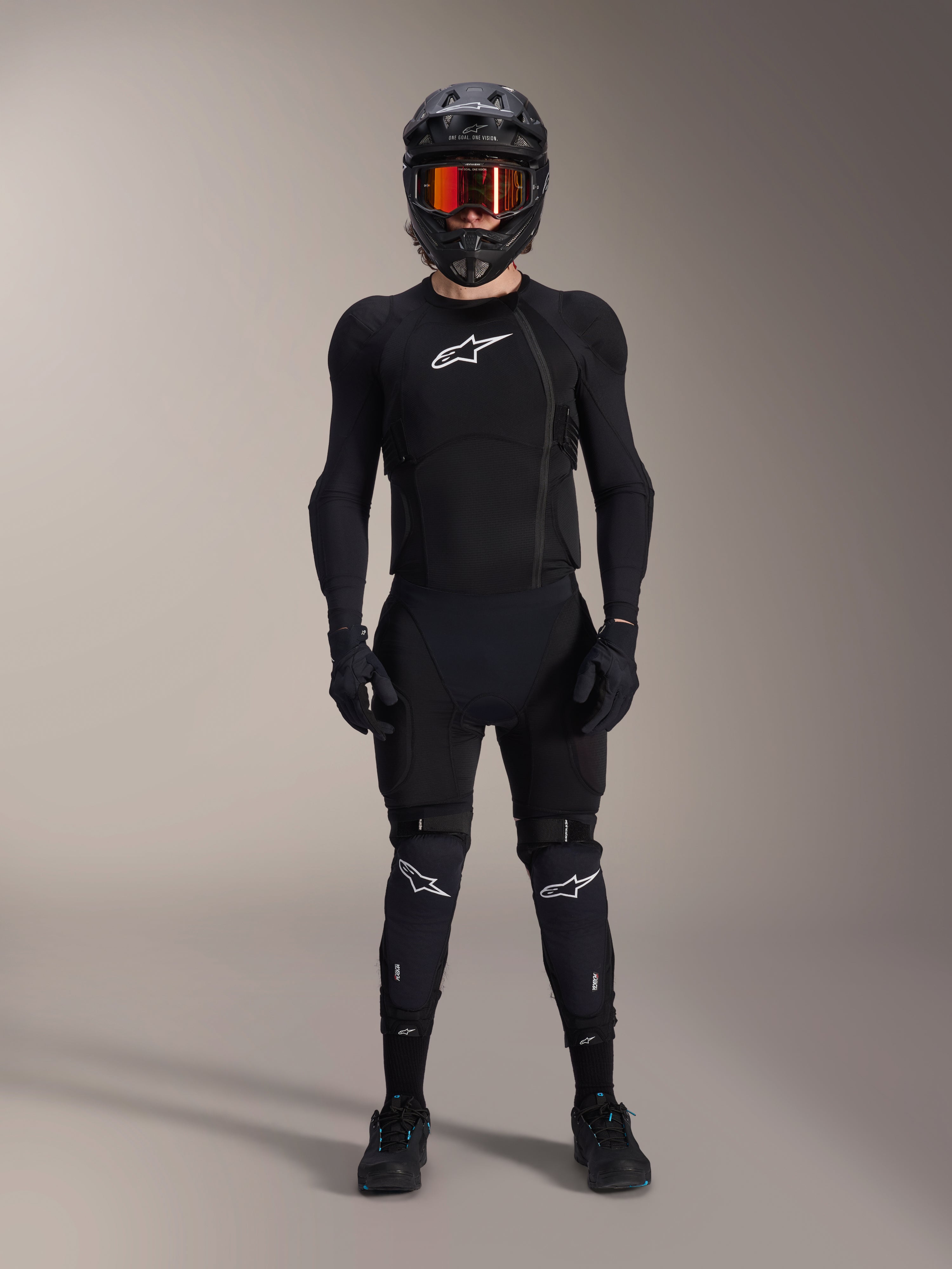 Mountain bike rider wearing complete protective gear, Alpinestars A-Impact Plasma Protection Jacket in Black and White, featuring a white chest logo and mesh fabric, worn with a black helmet, red-tinted goggles, and knee guards, coordinated high-performance MTB armor set.