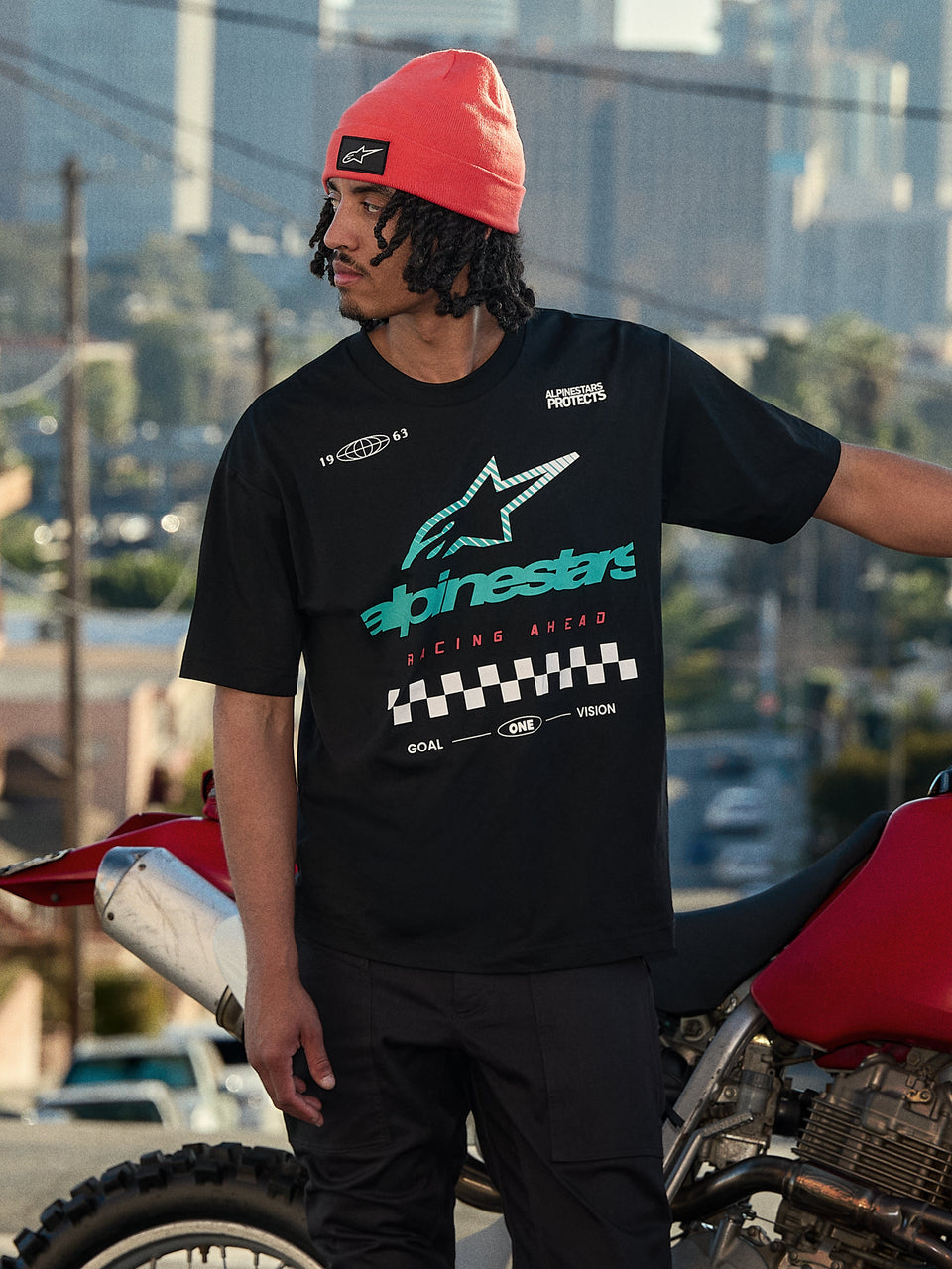 Alpinestars Aplomb Oversized CSF Tee, Short Sleeve T-Shirt, Black, featuring a large teal and white color-gradient Astars logo and checkered flag graphic, worn with a coral beanie, standing next to a red motorcycle, casual lifestyle streetwear