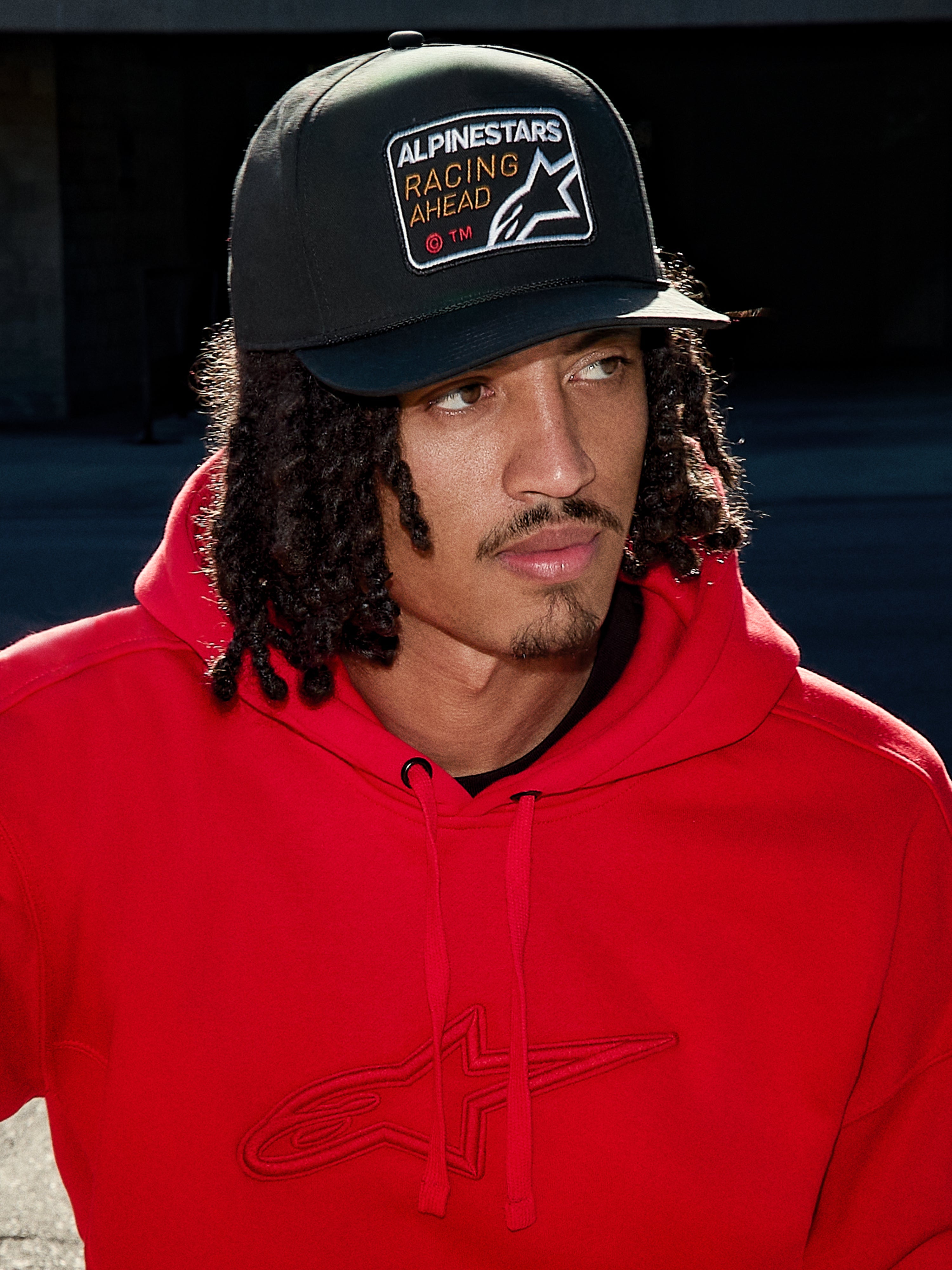 Mann mit Alpinestars Intuitive Snapback-Kappe in Schwarz, mit strukturierter Krone und 'Racing Ahead' Logo-Stickerei-Patch, kombiniert mit einem leuchtend roten Hoodie, lässige Lifestyle-Streetwear