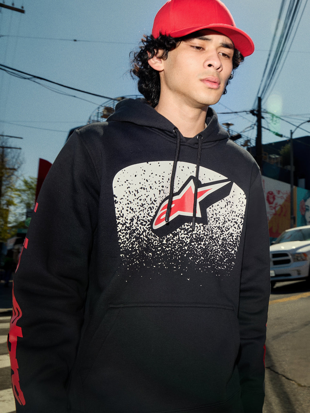 Alpinestars Obsessed Hoodie, schwarzer Fleece-Kapuzenpullover, weiße MX-Farbverlaufsgrafik auf der Brust mit rotem Stern-Logo, rote Wortmarken-Logos auf den Ärmeln, lässige, vom Motorsport inspirierte Streetwear in einer städtischen Umgebung