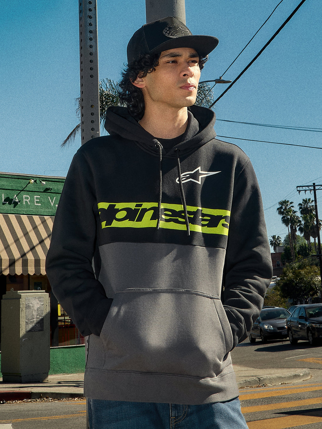 Alpinestars Parallel Hoodie, Lifestyle-Streetwear, Color-Block-Design in Schwarz und Gunmetal, limegrüner Bruststreifen mit Alpinestars-Logo, weißes Astars-Logo, Kapuze mit Kordelzug, getragen von einem Mann in einer urbanen Umgebung.