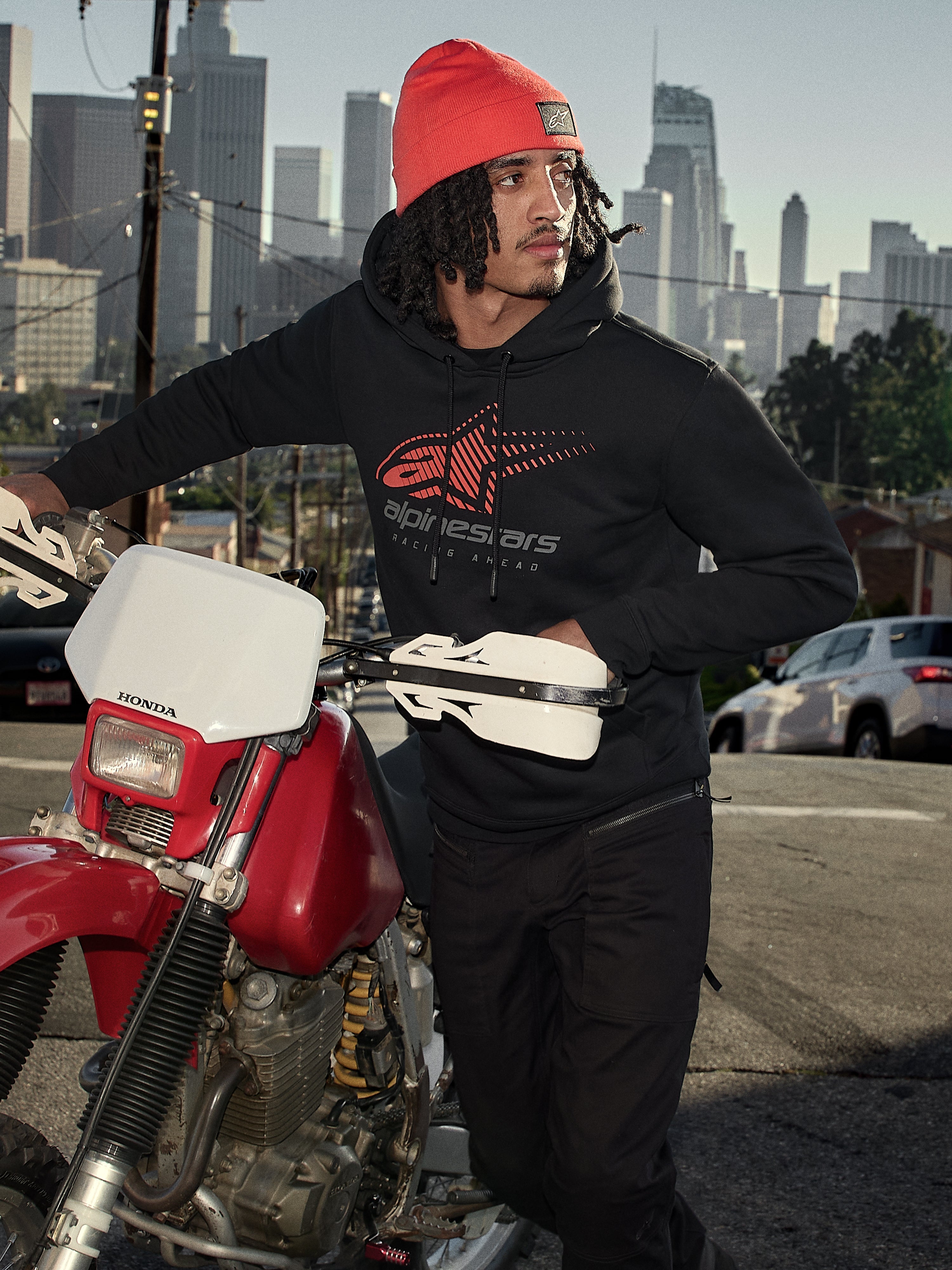 Alpinestars Pulse Hoodie, Black, hooded pullover fleece with red and grey Astars Combo Logo, worn by a man in an orange beanie standing next to a red Honda motorcycle in a city setting, casual motorsport streetwear