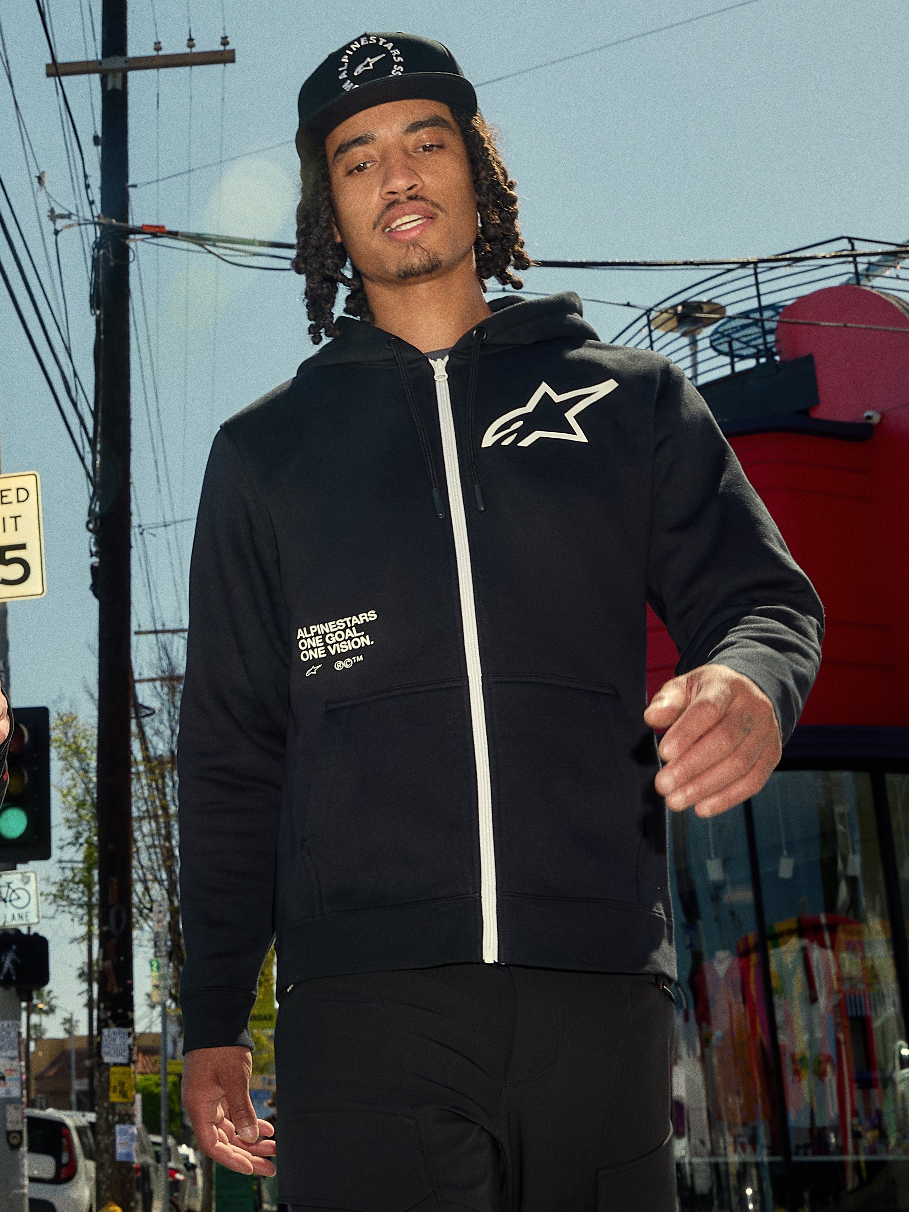 Alpinestars Transverse Hoodie, schwarzes Kapuzenfleece mit Reißverschluss vorne, weißer Reißverschluss und weißes Astars-Logo auf der Brust, „One Goal One Vision“ Textaufdruck, lässiger Streetwear-Stil, getragen von einem Mann in einer urbanen Außenumgebung
