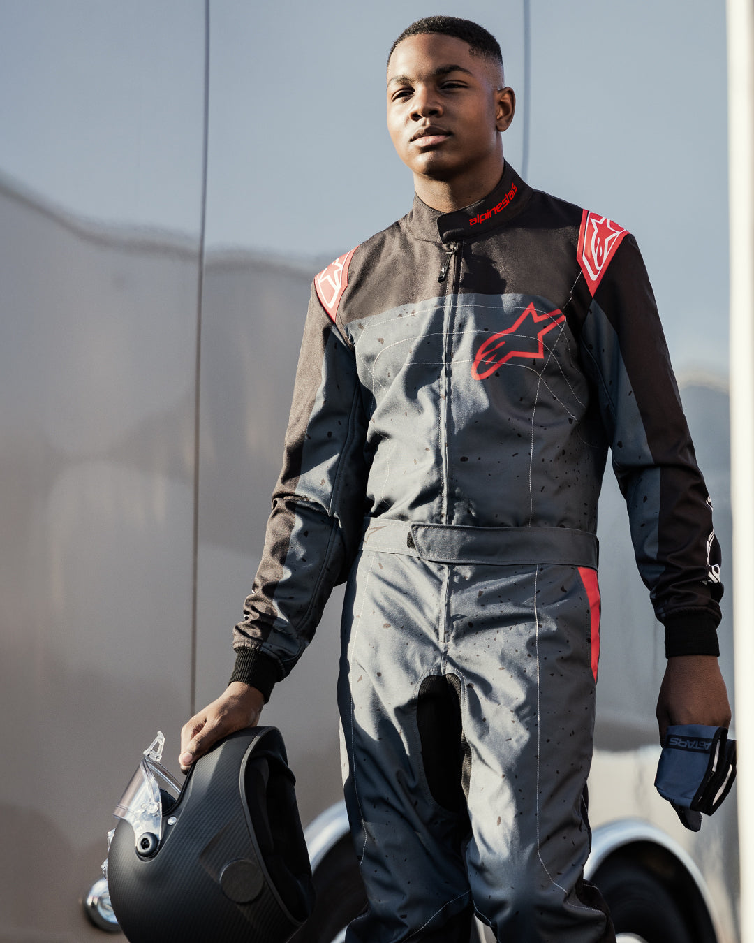 Kartfahrer im Alpinestars KMX-9 V3 Graphic 6 Anzug in Anthrazit-Schwarz und Rot, hält einen mattschwarzen Helm und Handschuhe, mit markanten roten Alpinestars-Logos und gesprenkeltem Grafikdesign, Outdoor-Motorsport-Umfeld.