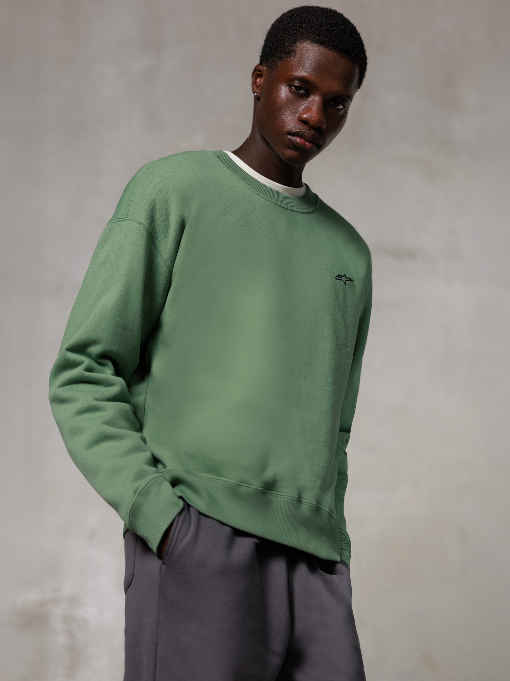 Alpinestars Essentials Ageless Crew, Rundhals-Sweatshirt, Kaktusgrün, mit einem kleinen schwarzen Alpinestars-Logo auf der Brust, getragen von einem Model mit grauer Hose, lässige Lifestyle-Streetwear
