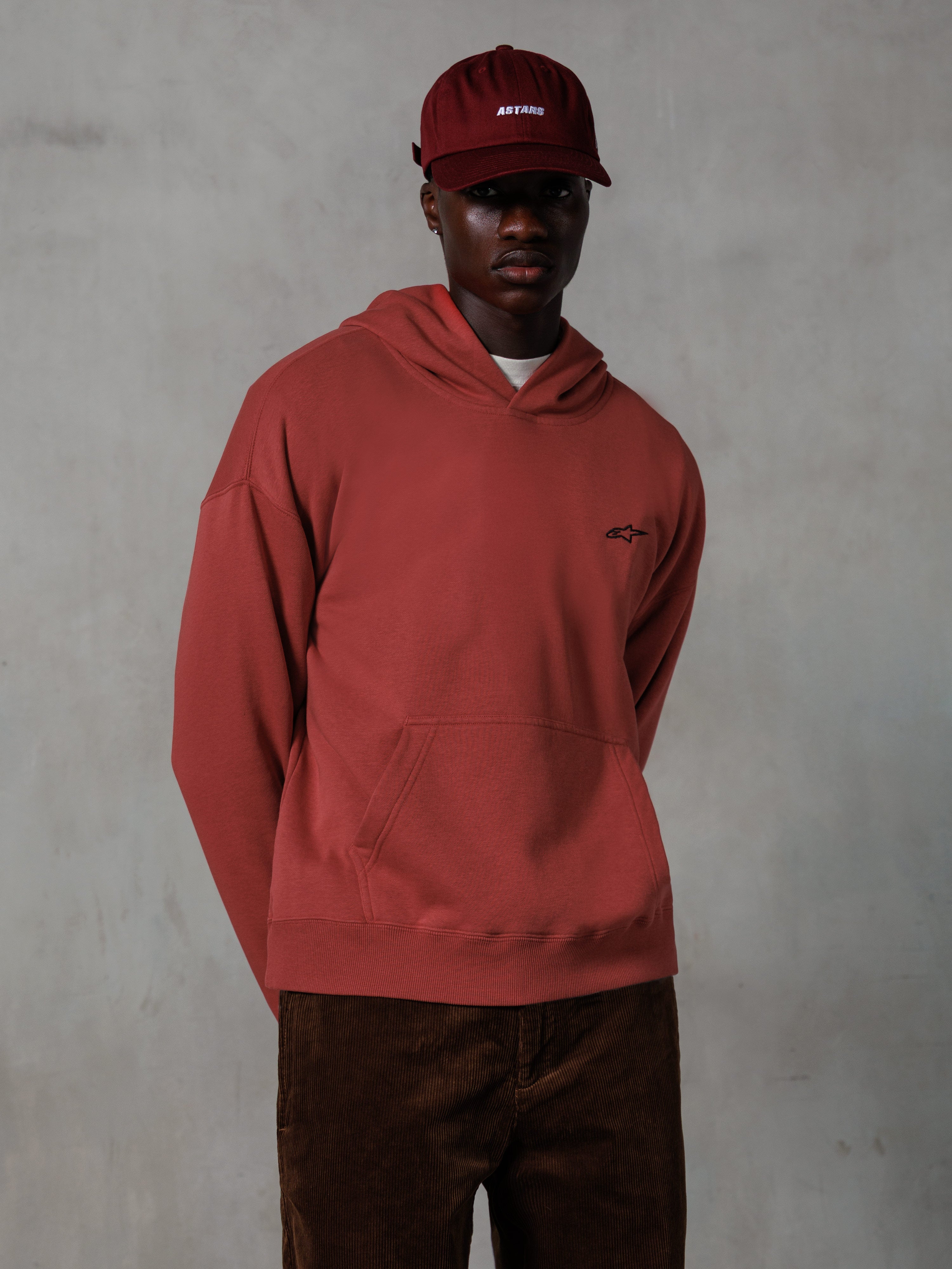 Alpinestars Essentials Ageless Hoodie, lässige Streetwear, Ziegelrot, kleines schwarzes Brustlogo, Kängurutasche vorne, getragen mit passender roter ASTARS Kappe und brauner Cordhose, schlichtes minimalistisches Design