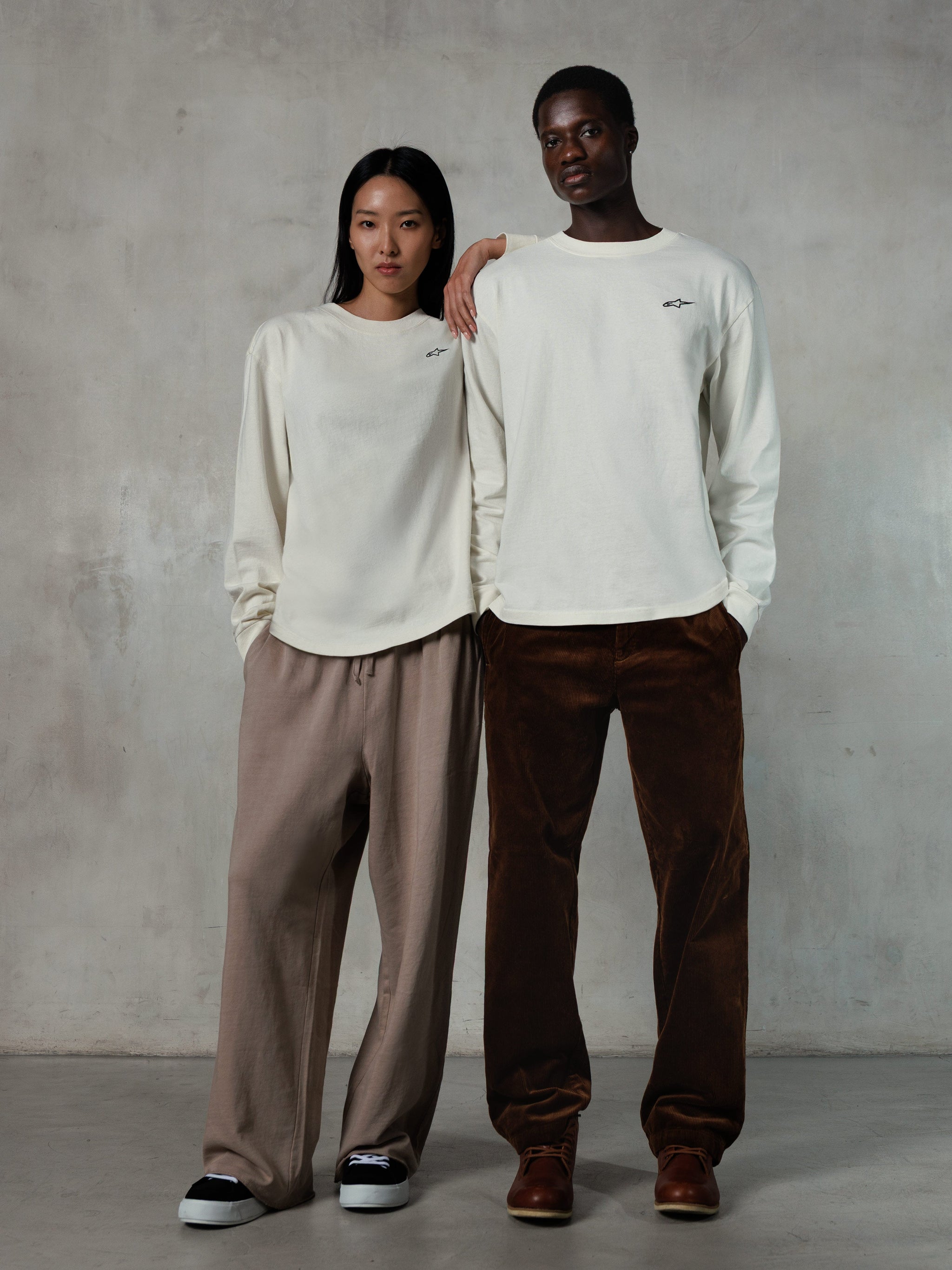 Alpinestars Essentials Ageless LS Tee, Long-Sleeve T-Shirt, Off White, worn by male and female models, small black Alpinestars logo on the chest, relaxed fit, styled with tan sweatpants and brown corduroy pants, casual motorsport-inspired streetwear
