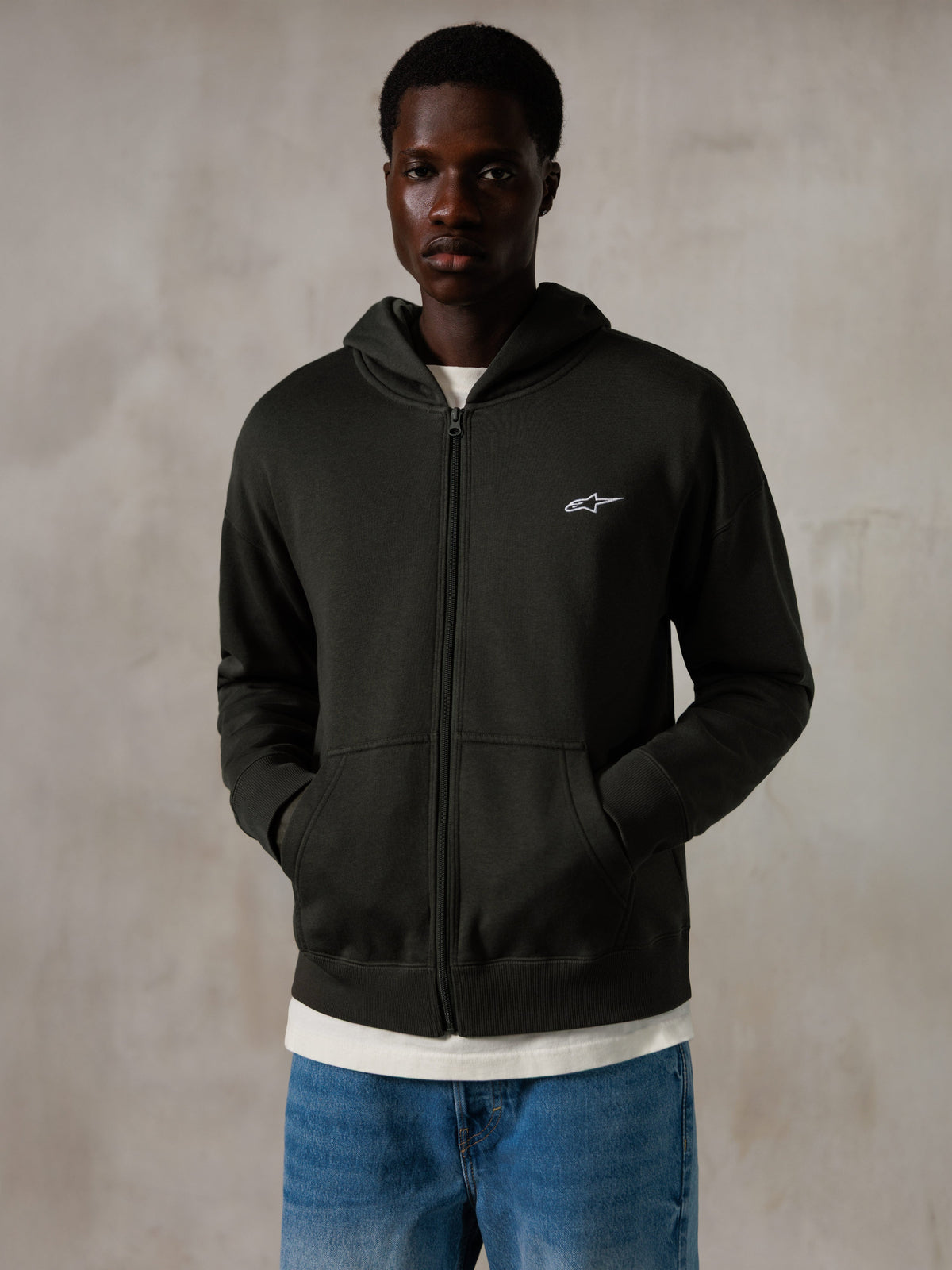 Alpinestars Essentials Ageless Zip Hoodie, Off Black, Zip-Kapuzenjacke mit einem kleinen weißen Alpinestars-Logo auf der Brust, entspannte Passform, getragen von einem Modell mit blauen Jeans und einem weißen T-Shirt, lässige vom Motorsport inspirierte Streetwear