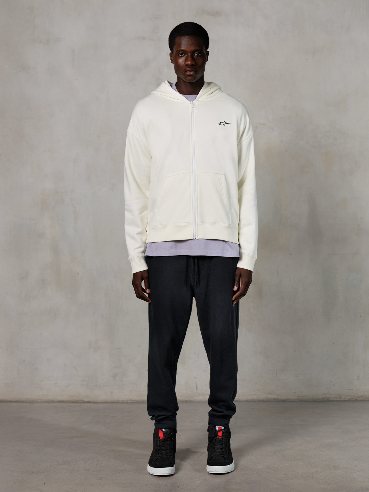 Alpinestars Essentials Ageless Zip Hoodie, lässige Streetwear, Off-White mit dezentem schwarzem Logo auf der Brust, durchgehender Reißverschluss vorne, Kapuzendesign, entspannte Passform, kombiniert mit schwarzer Jogginghose und Sneakern