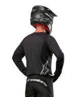 Alpinestars 2024 Fluid Graphite Ausrüstungsset, Motocross-Trikot und -Hose, Graphit und Silber, Rückansicht mit silbernem Branding am unteren Rücken und an der Taille, getragen mit schwarzem Helm in Carbon-Optik, leichtes Poly-Gewebe-Design für Offroad-Fahrten