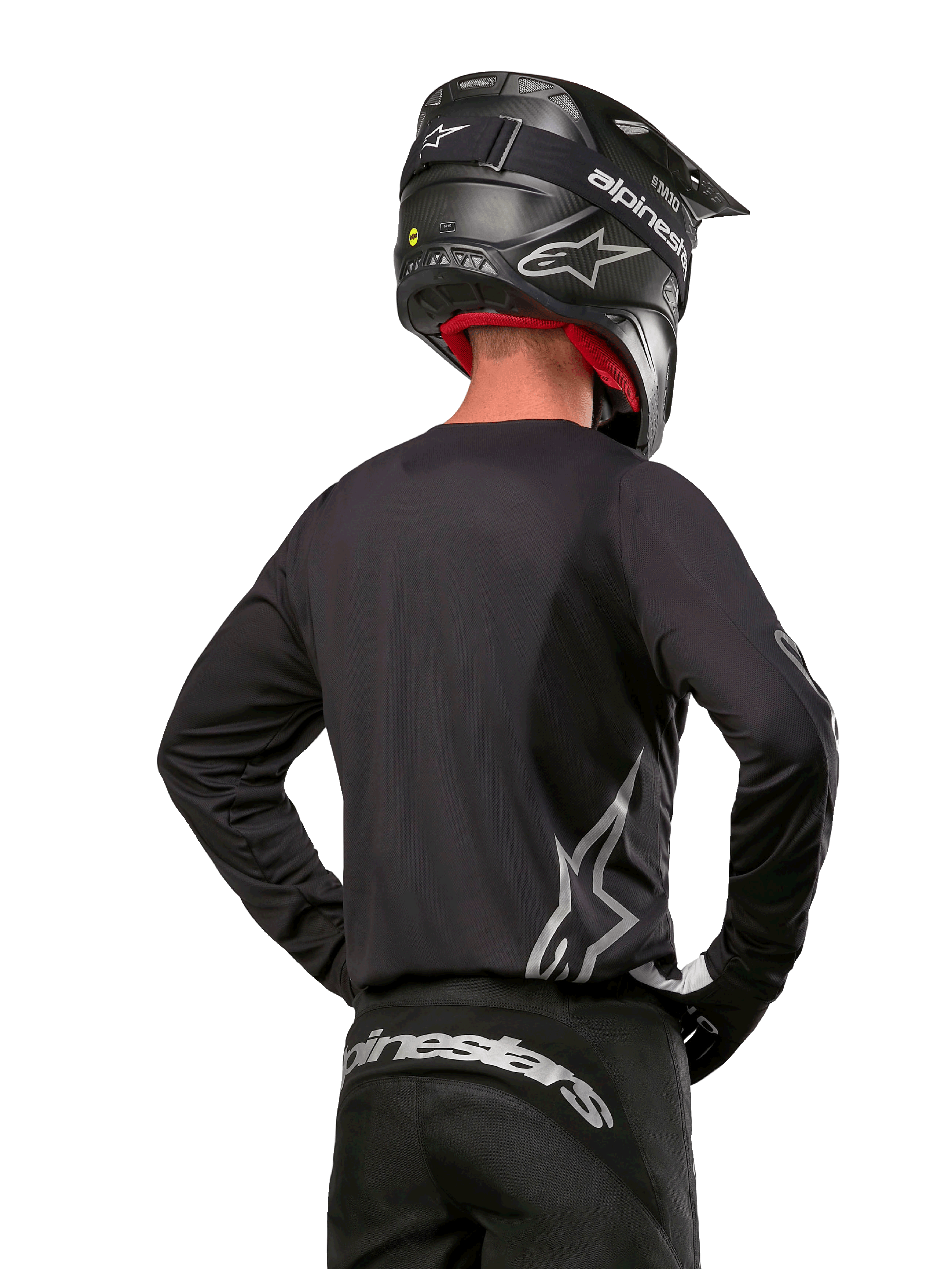 Motocross rider viewed from the back wearing the Alpinestars 2024 Fluid Graphite Jersey in Black Silver, featuring a large silver logo on the side, paired with a black carbon-texture helmet and black pants, professional off-road racing gear