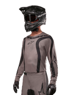 Alpinestars 2024 Fluid Lurv Trikot, Motocross-Jersey, Mud Black und Grau, langärmliges Design mit geschwungenen schwarzen Streifen und Logos, getragen mit passender Hose und einem schwarzen Motocross-Helm, abgestimmte Offroad-Rennausrüstung