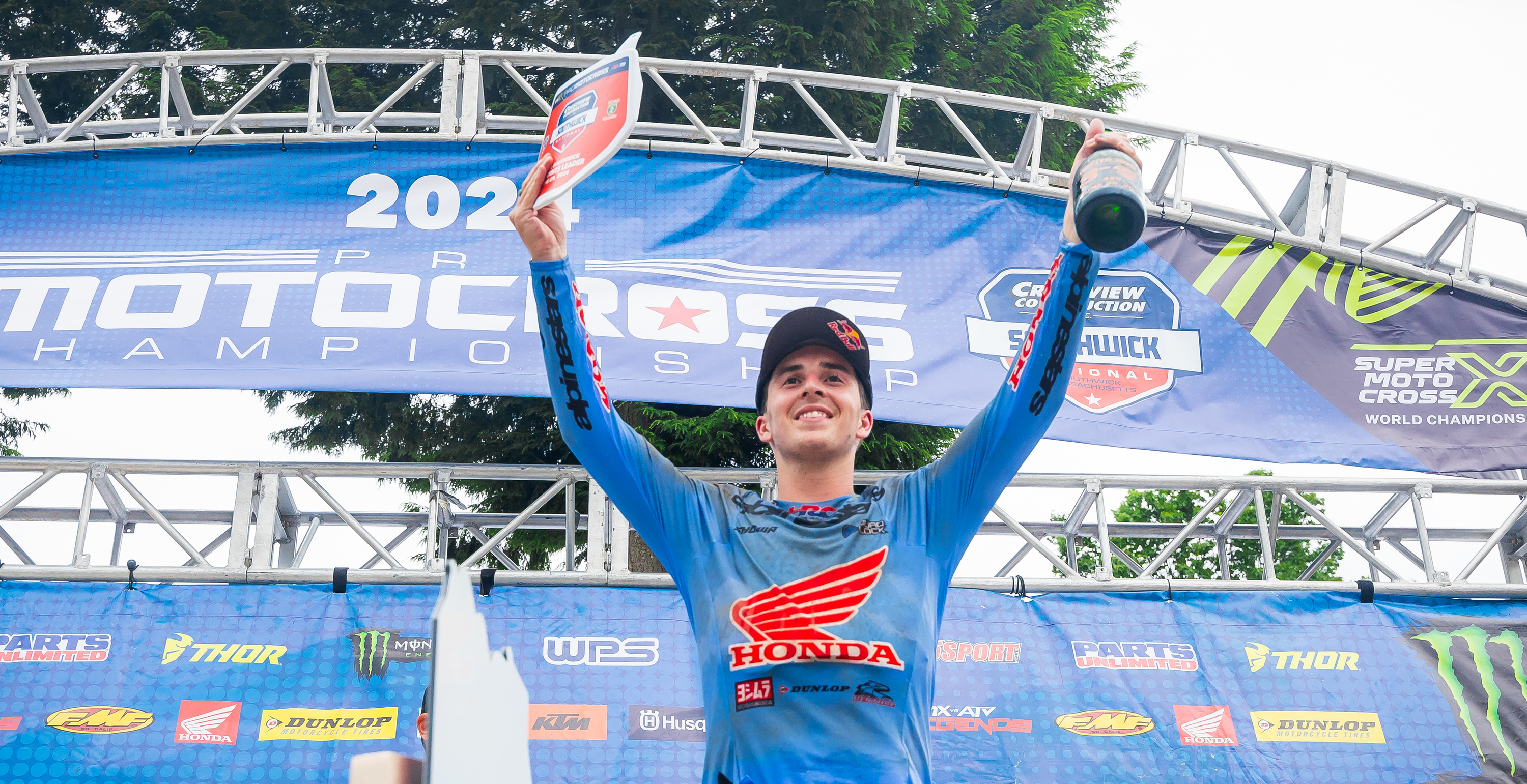 Hunter Lawrence celebrating on a podium with a trophy at a motocross event, surrounded by sponsor banners.