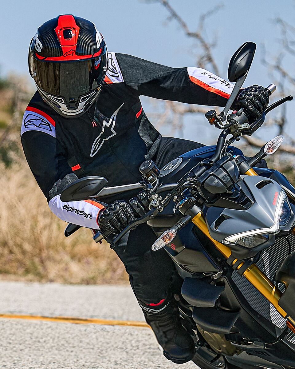 Motorradfahrer in Schräglage auf einem Naked Sportbike, trägt die Alpinestars T-SPS Air v2 Jacke in Schwarz mit weißen und neonroten Akzenten, kombiniert mit einem schwarz-roten Helm und schwarzen Handschuhen, leistungsorientierte Textilausrüstung für urbanes sportliches Fahren.