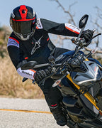 Motorradfahrer in Schräglage auf einem Naked Sportbike, trägt die Alpinestars T-SPS Air v2 Jacke in Schwarz mit weißen und neonroten Akzenten, kombiniert mit einem schwarz-roten Helm und schwarzen Handschuhen, leistungsorientierte Textilausrüstung für urbanes sportliches Fahren.