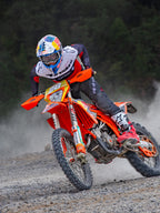 Motocross-Fahrer in Aktion auf einem orangefarbenen KTM-Bike, trägt Alpinestars Pro-Dura Ausrüstung bestehend aus einem rot-weiß-schwarzen Trikot, einer dunkelgrauen Hose, rot-weißen Stiefeln und einem Red Bull Rennhelm, während einer scharfen Kurve auf einem Schotterweg