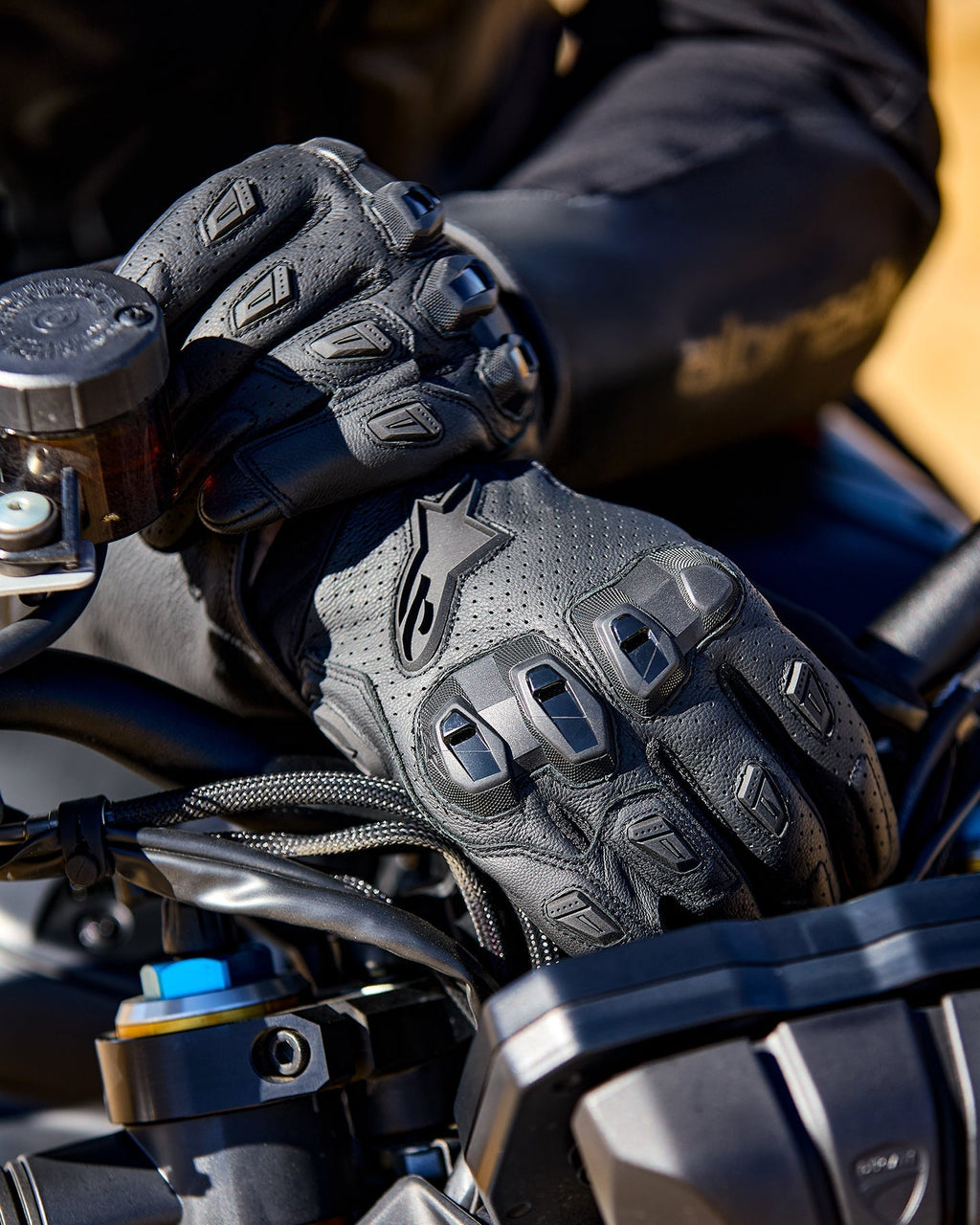 Alpinestars SP X 7 Gloves, black perforated leather motorcycle gloves featuring TPU knuckle protectors and finger sliders, shown on a rider's hands gripping motorcycle handlebars.