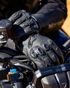 Alpinestars SP X 7 Gloves, black perforated leather motorcycle gloves featuring TPU knuckle protectors and finger sliders, shown on a rider's hands gripping motorcycle handlebars.