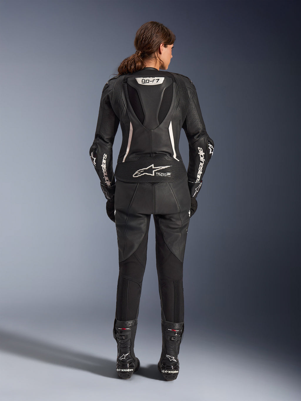 Alpinestars Stella GP-R7 2-Piece Leather Suit, Women's Motorcycle Suit, Black with white accents, rear view showing GP-R7 aerodynamic hump and Tech-Air Ready branding, white Alpinestars logos on sleeves, accordion stretch panels, worn with black racing boots
