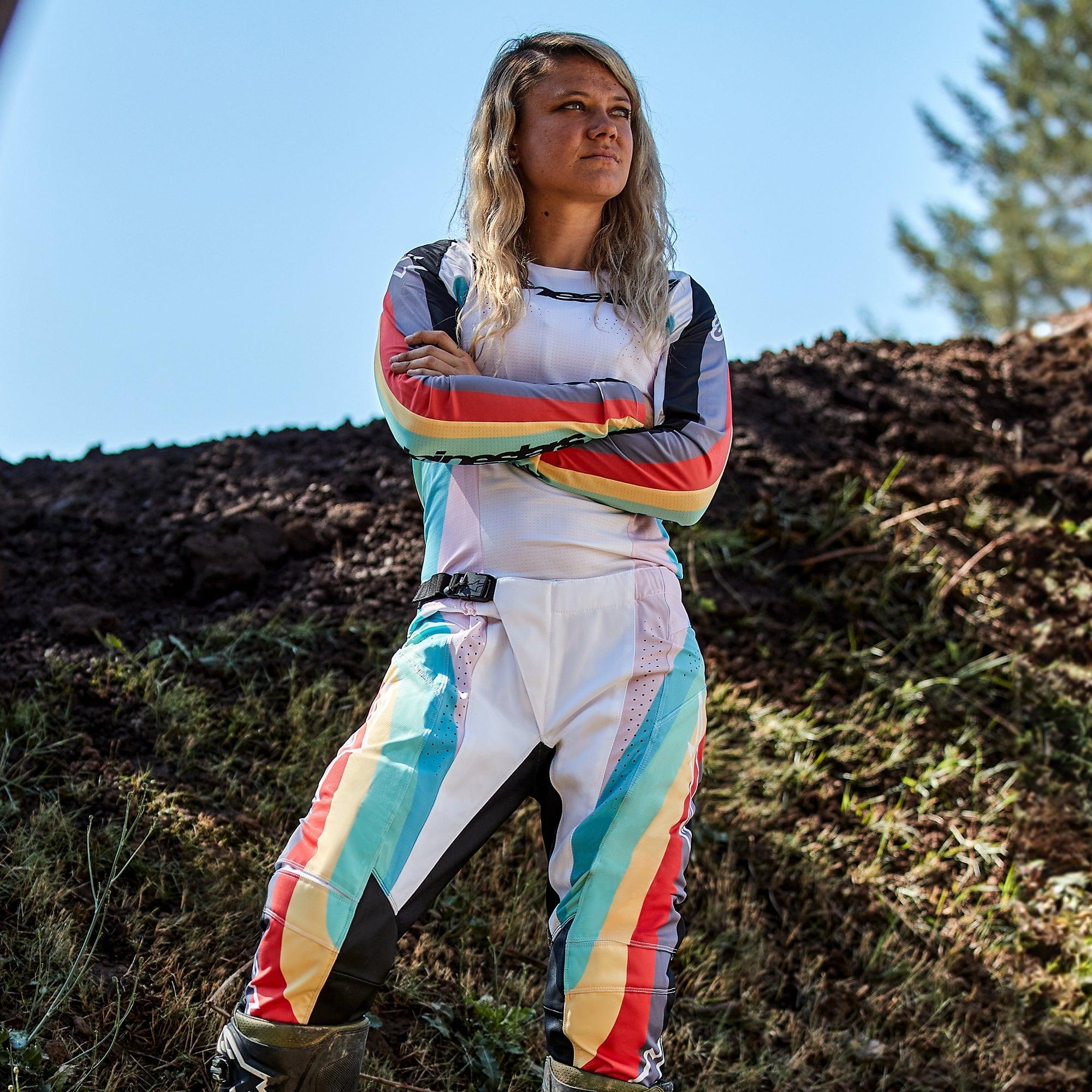 Female motocross rider wearing Alpinestars Women Stella 2024 Techstar Pants and matching jersey, Black Multicolor design with white base and vibrant blue, yellow, and red stripes, standing with arms crossed on a dirt track, professional off-road racing outfit