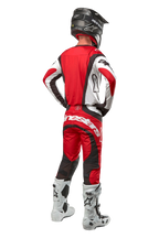 Alpinestars 2024 Techstar Ocuri Hose, Motocross-Hose, Mars Rot Weiß Schwarz, Rückansicht eines Motocross-Fahrers in kompletter Rennausrüstung, einschließlich passendem Jersey, schwarzem Helm und weiß-schwarzen Stiefeln, hebt das Alpinestars-Branding und die strapazierfähige Stoffkonstruktion hervor.
