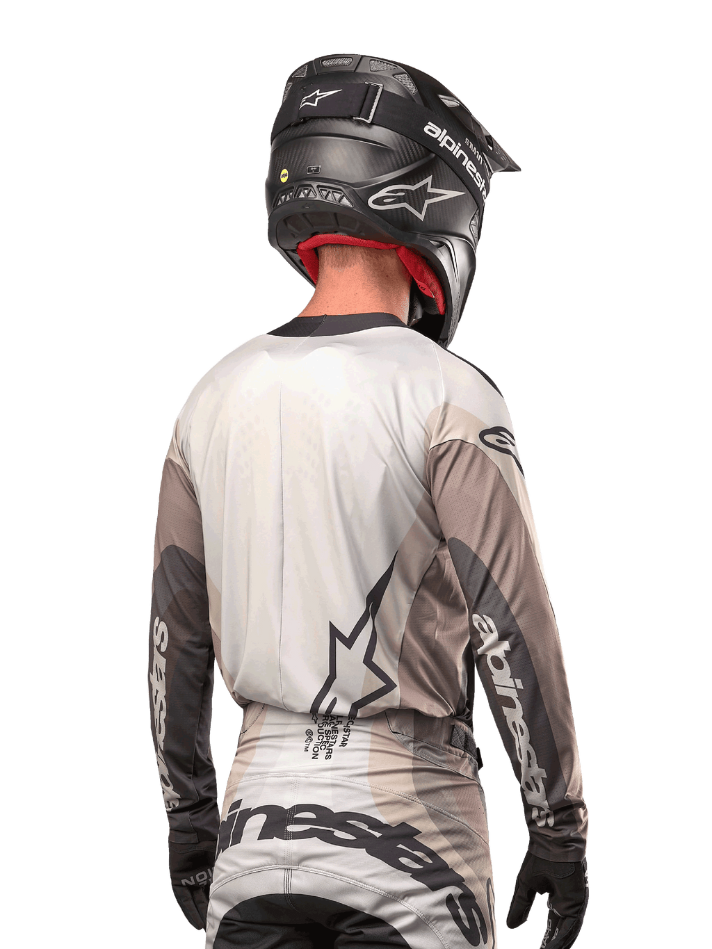 Motocross rider from behind wearing the Alpinestars 2024 Techstar Pneuma Jersey in Dark Sand Iron Dust Gray, paired with a black carbon-finish helmet, featuring a large star logo on the back and Alpinestars branding on the sleeves.