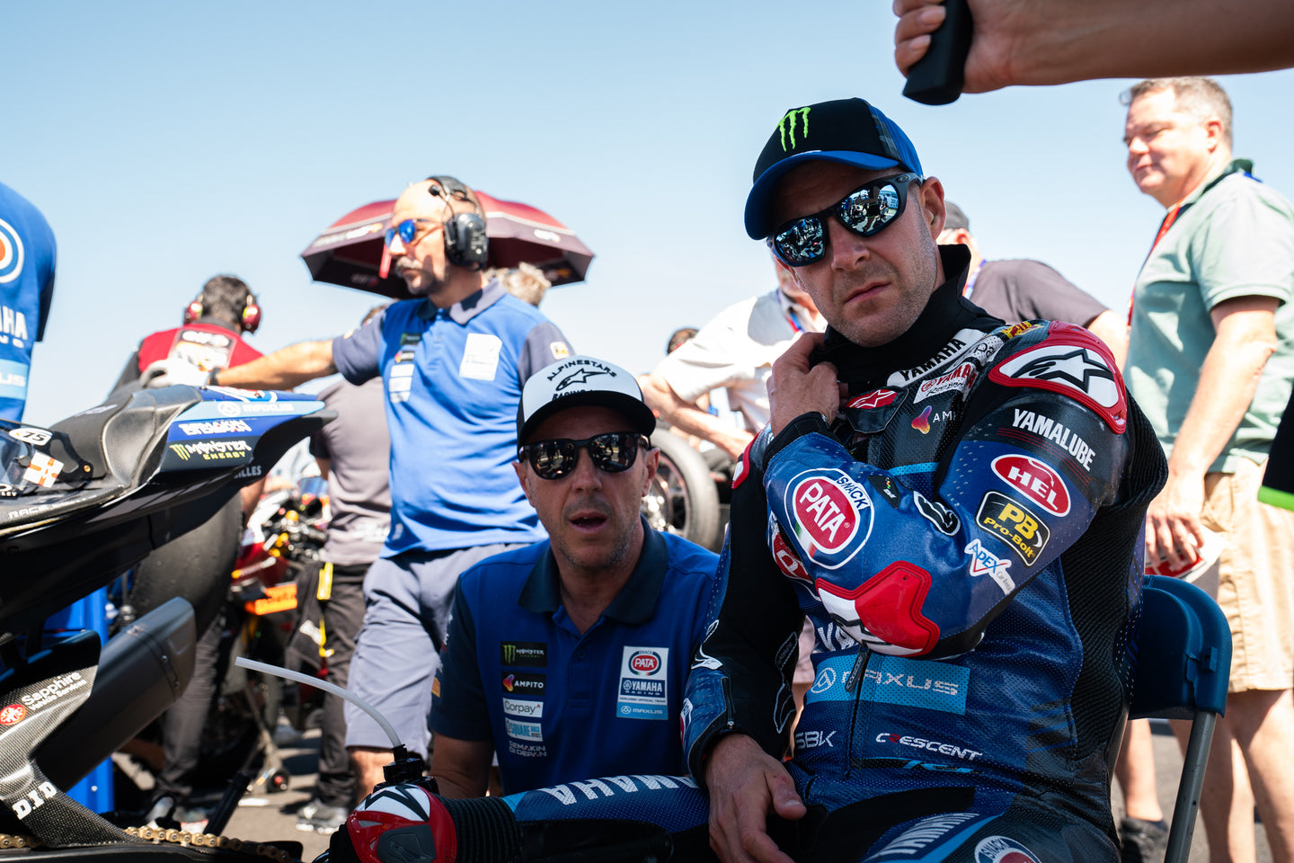 Jonathan Rea in a colorful Alpinestars suit with sponsor logos sitting on a motorcycle, surrounded by people and racing equipment.