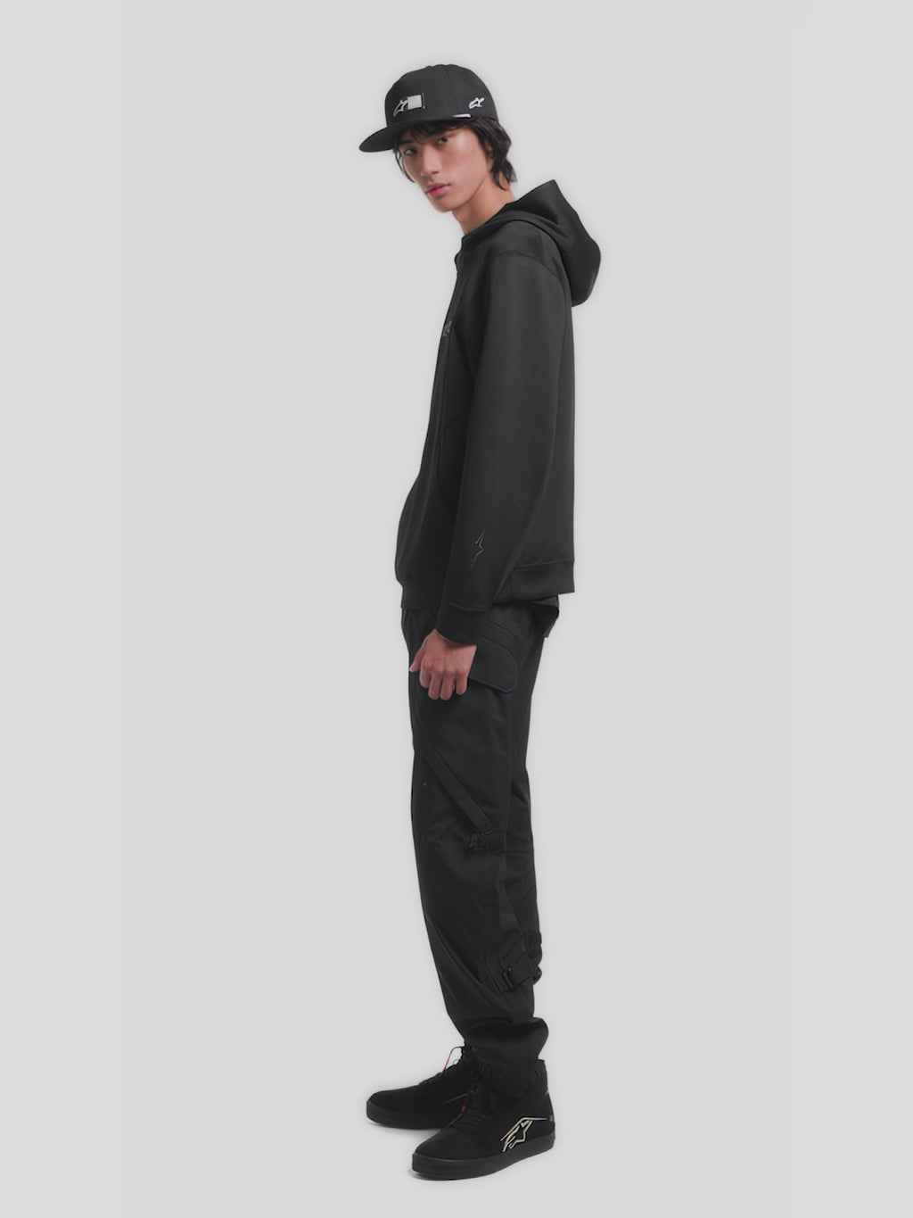 Alpinestars Label Snapback Hat, Black Sportswear Cap for Men and Women, featuring a flat bill, structured crown, woven front label, and white embroidered side logo. Model shown wearing the hat, highlighting the adjustable snapback closure and premium twill construction.